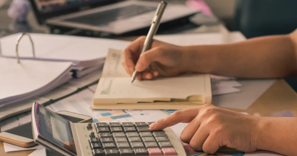 Hands using a calculator and writing in a planner while working on payroll for small business records